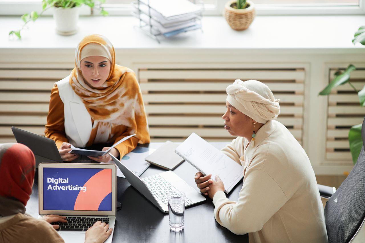 services-04 Three women in hijabs collaborating on digital advertising strategies around a table with laptops.