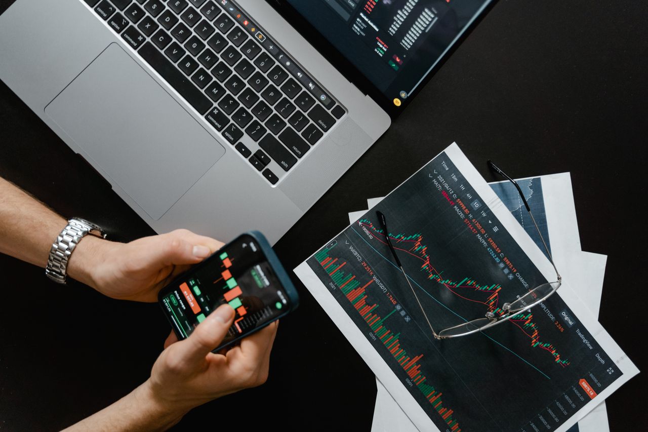 about-02 Hands using smartphone beside laptop with stock charts, showcasing digital trading.